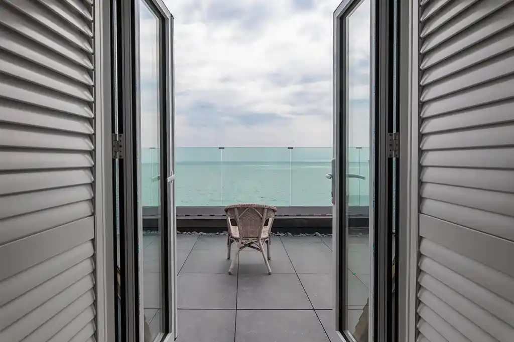 View through open plantation shutters onto a private tiled balcony with a single wicker chair and a glass balustrade overlooking the ocean at Selsey Beach House.