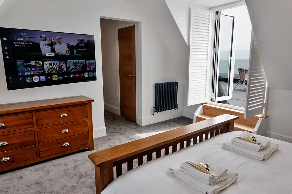 Bedroom at Selsey Beach House featuring a wooden bed, a large wall-mounted smart TV, and steps leading out to a private balcony.