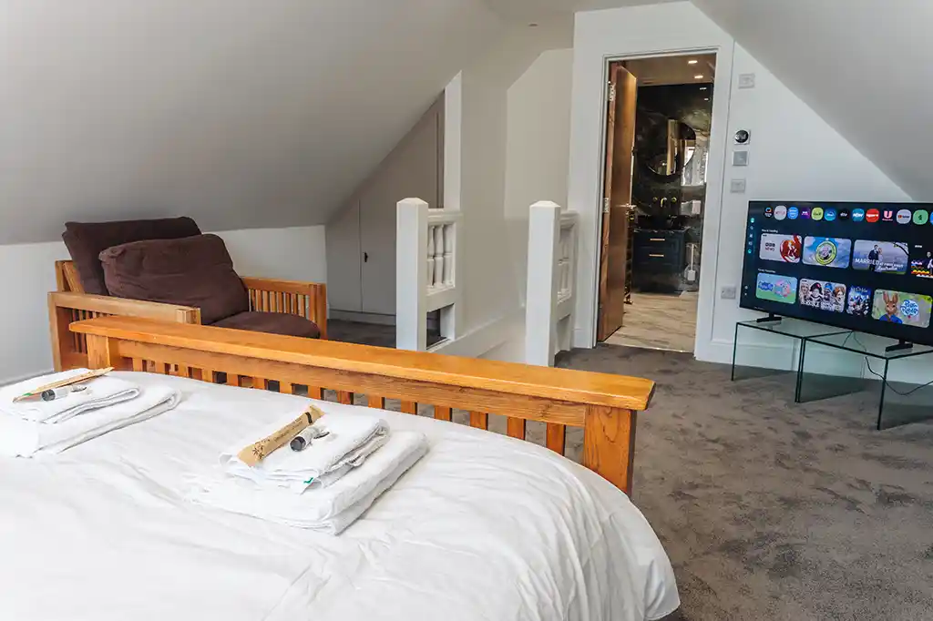 Spacious bedroom at Selsey Beach House featuring a wooden bed, a large flat-screen smart TV, and an open doorway leading to a modern en-suite bathroom.