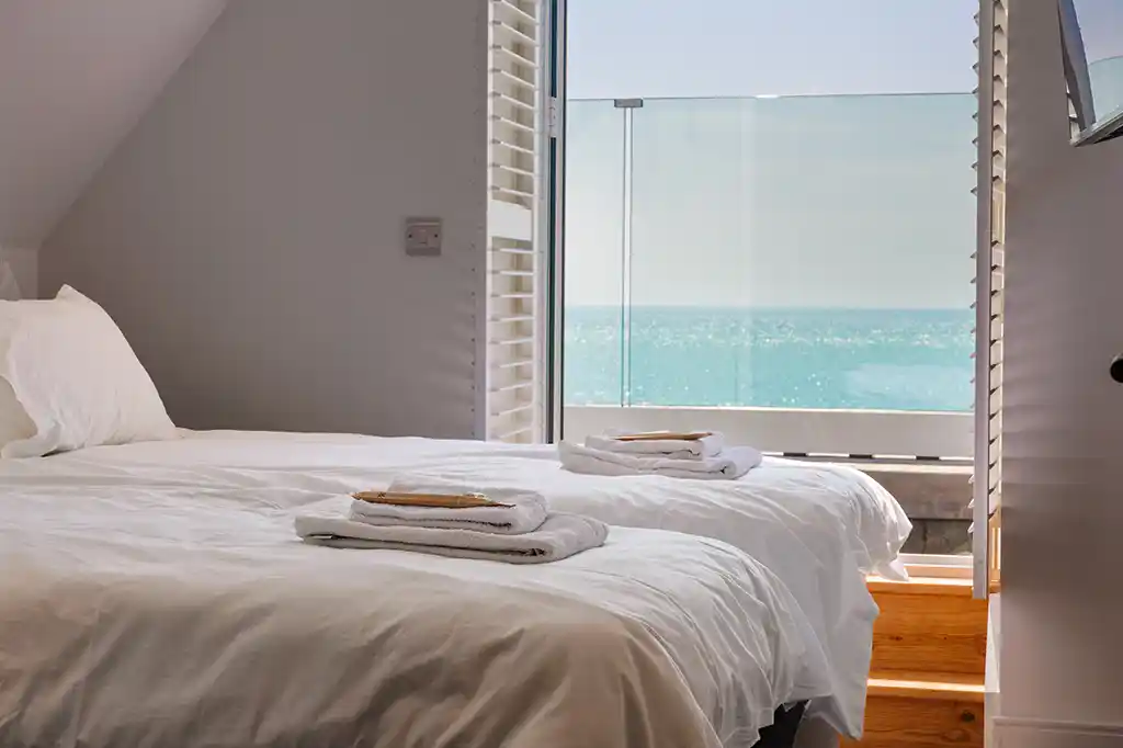Bright bedroom at Selsey Beach House featuring two single beds with fresh white linens and open shutters framing a beautiful ocean view.