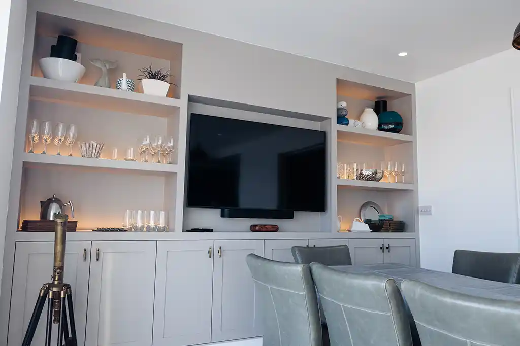 Modern dining room at Selsey Beach House featuring a built-in media wall with a large flat-screen TV, illuminated shelving, and grey dining chairs.