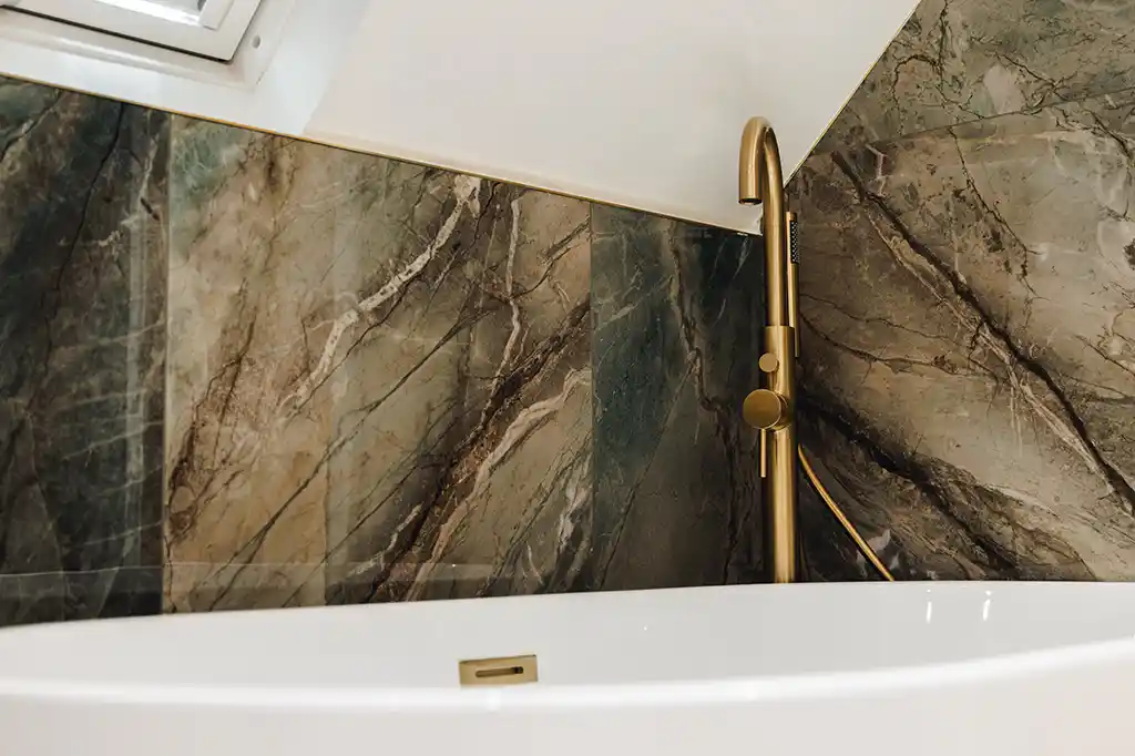 Close-up of a freestanding white bathtub and modern gold floor-standing tap against green marble tiles in the luxury en-suite at Selsey Beach House.