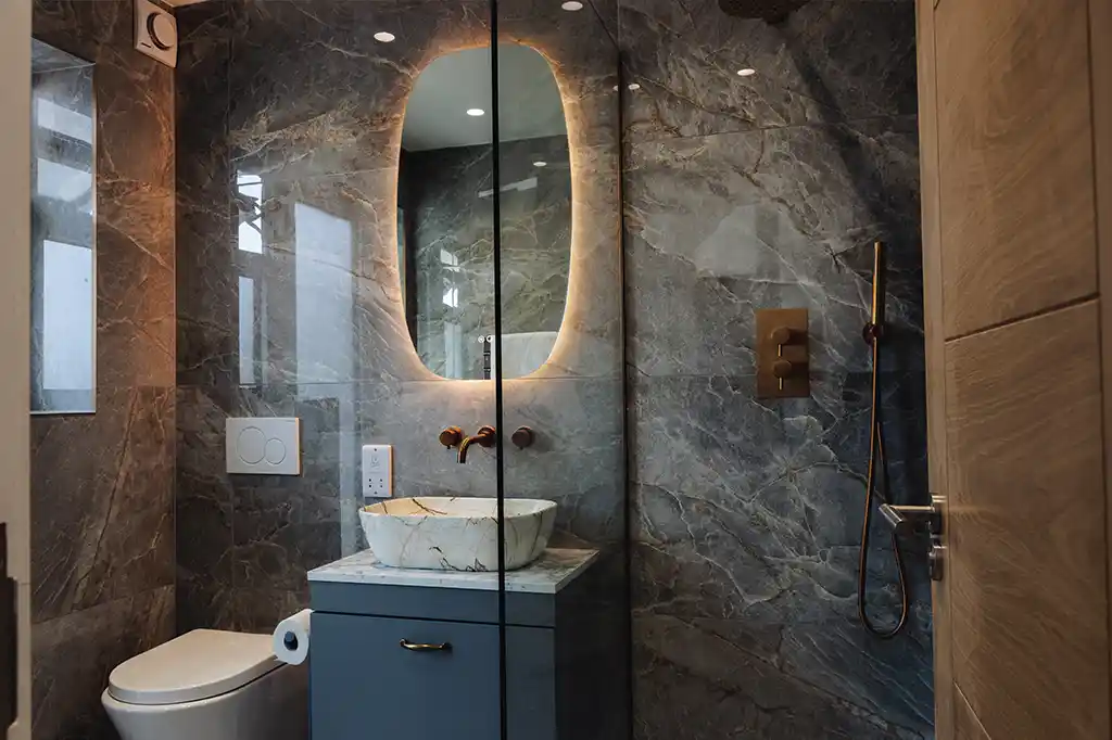 Luxury modern bathroom at Selsey Beach House featuring dark marble-effect tiles, a backlit oval mirror, and bronze fixtures.