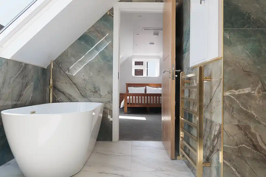 Luxury en-suite bathroom at Selsey Beach House featuring a freestanding white bathtub, green marble tiles, and a gold heated towel rail.
