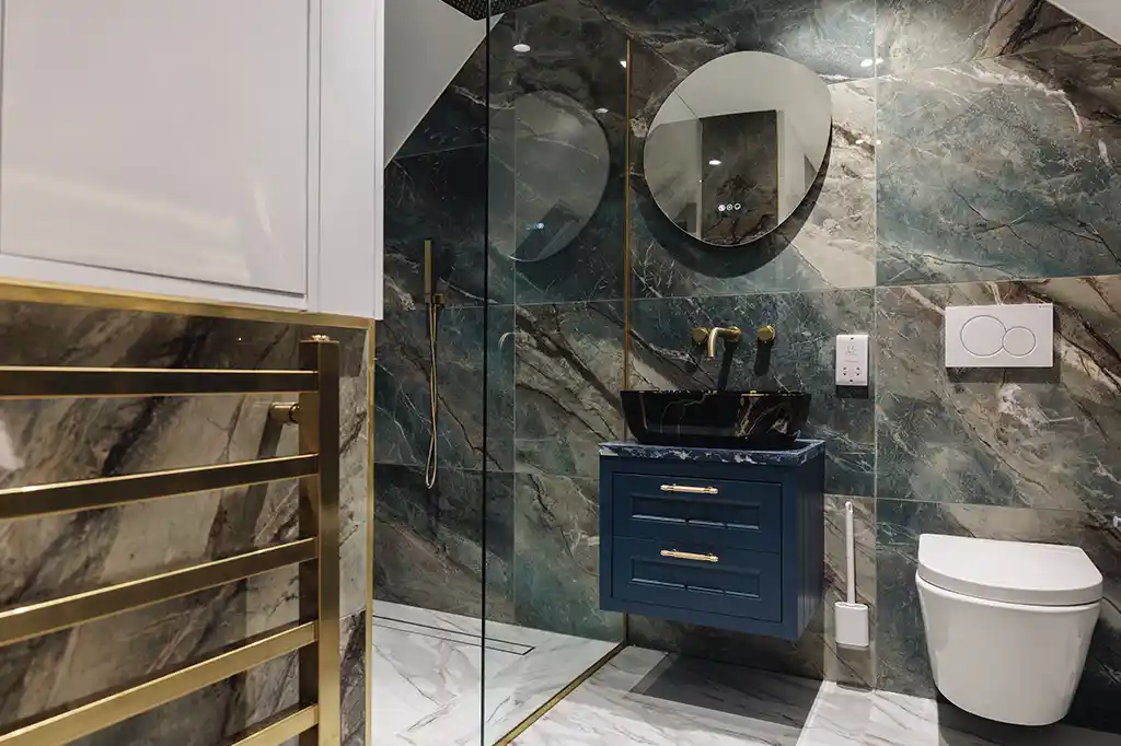 Opulent modern bathroom at Selsey Beach House featuring green marble-effect tiles, a floating blue vanity with a black sink, and elegant gold fixtures.