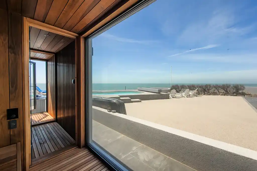 View from inside the outdoor wooden sauna at Selsey Beach House looking through a large panoramic window towards the patio, swimming pool, and ocean.
