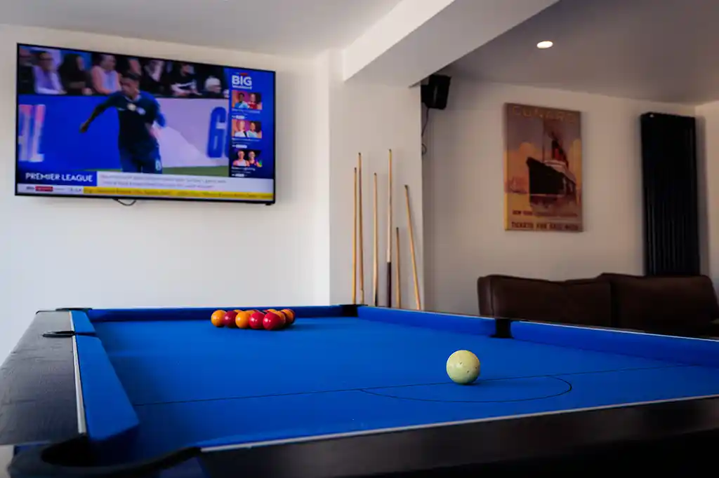 Close-up of a vibrant blue pool table ready for a game, with a large wall-mounted TV showing sports in the background at Selsey Beach House.