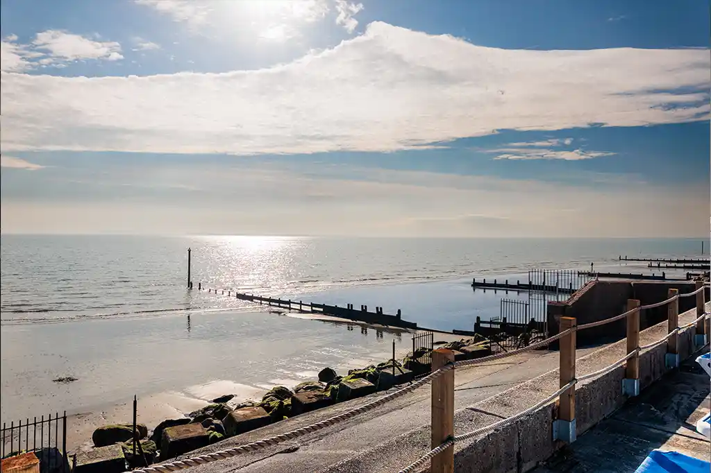 Ocean view from the garden at Selsey Beach House, showing a rope railing promenade, sandy beach, and wooden groynes.